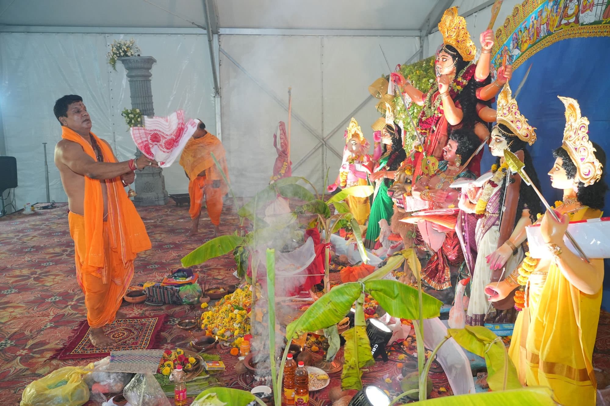 A priest in saffron robes waves an Assamese gamosa during the aarti before the Durga idol and her family, with banana plants, flowers and rising smoke in front