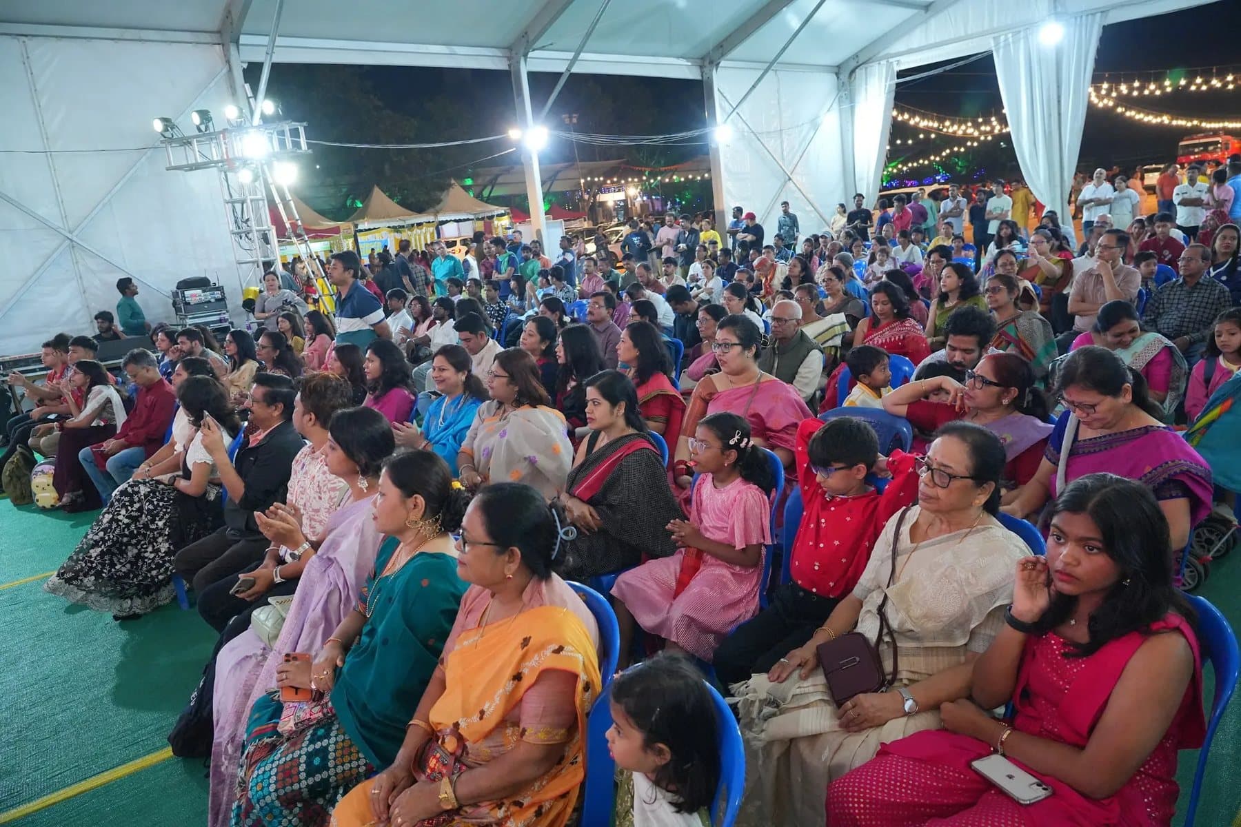 Families, elders and children seated on chairs under a large tent at night, watching the evening programme at the 2025 Durga Puja, with strings of lights behind them