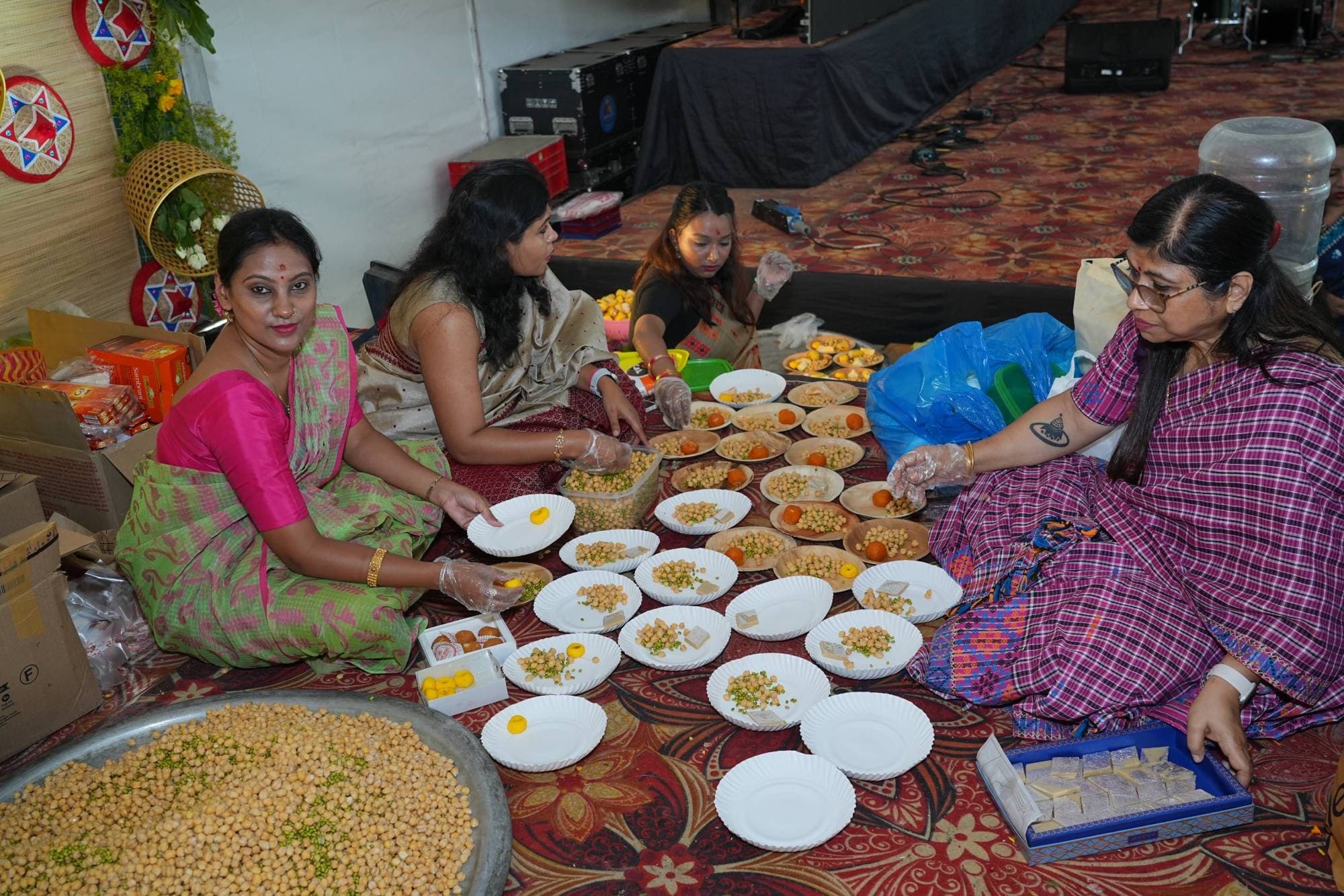 Volunteers in Assamese mekhela chador sitting on the floor, wearing gloves and arranging prasad of chickpeas, sweets and fruit onto plates