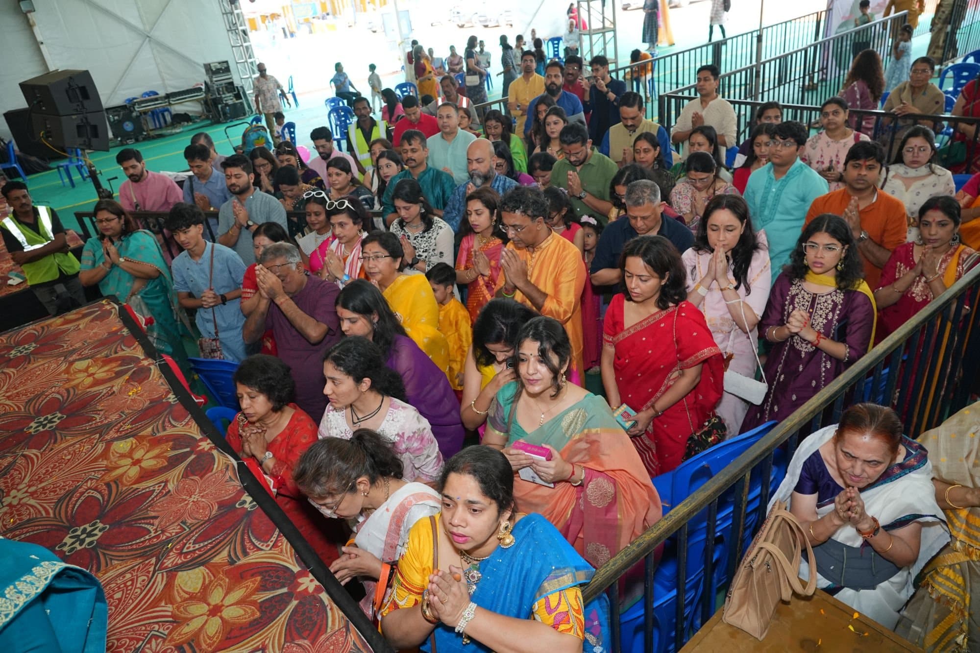 Devotees of all ages standing close together at the edge of the stage with folded hands, offering Pushpanjali at the 2025 Puja