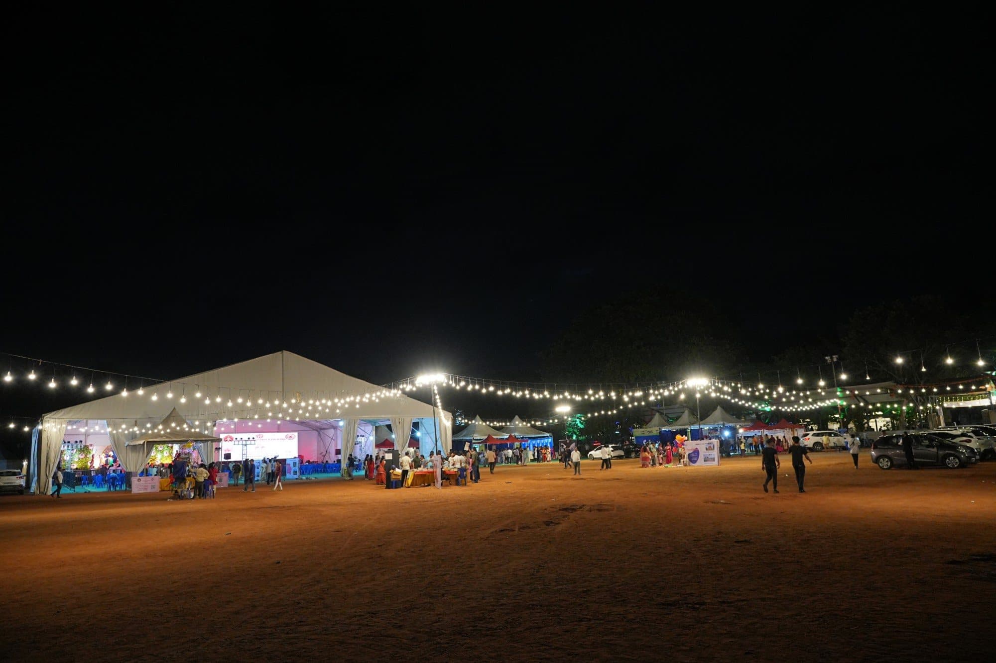 The Puja grounds at night: a large white tent hung with strings of lights, the stage glowing inside, stalls and families on the open ground